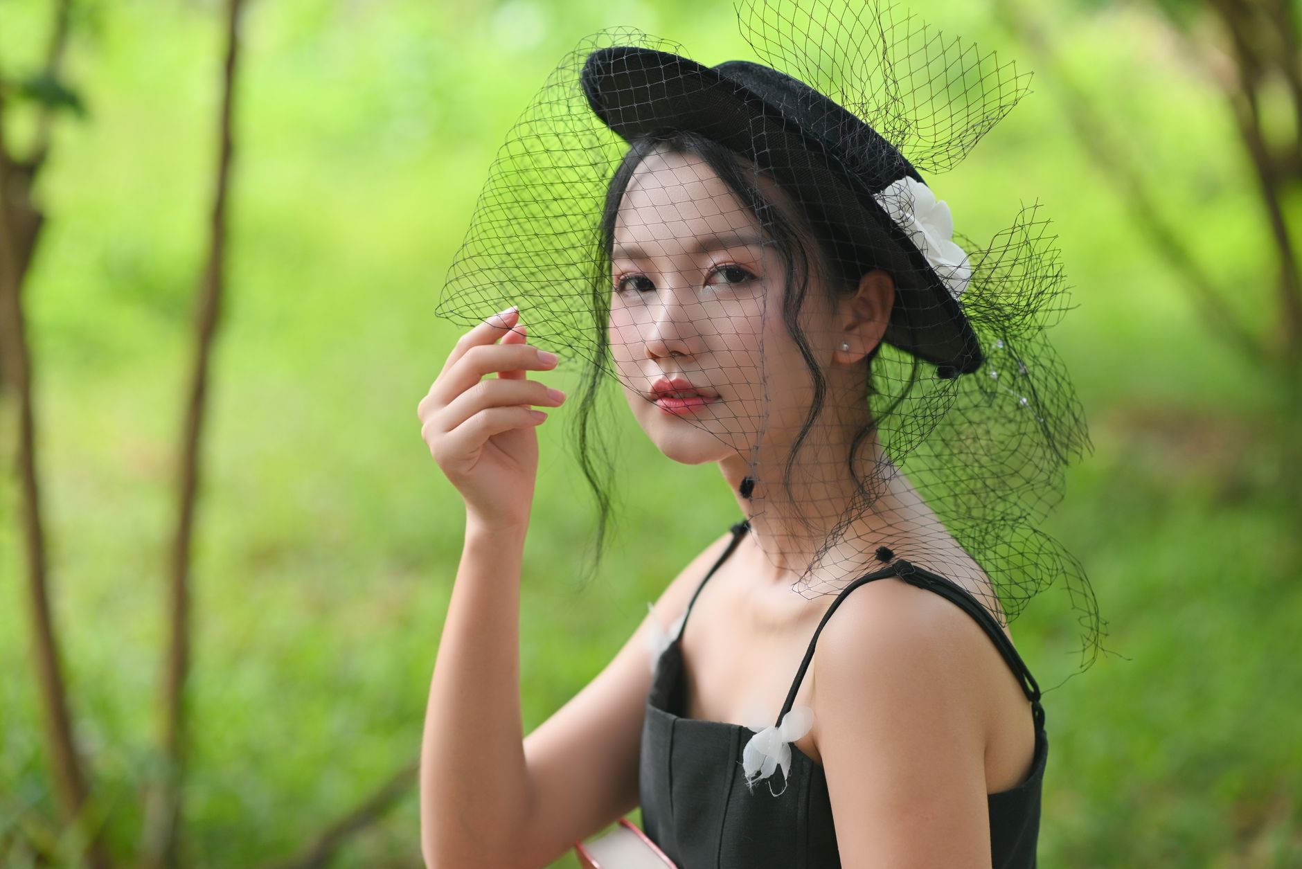 Stylish portrait of a woman wearing a black dress and veiled hat, set against a lush green outdoor background. — The Art of M